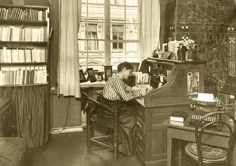 Ruskeansävyinen valokuva huoneesta, jossa nainen istuu kirjoituspöydän ääressä kirjoittamassa. Seinällä on kirjahylly, toisella seinällä kakluuni, etualalla pieni pöytä jolla on kirjoituskone. Ikkunasta näkyy vastapäisen kivitalon ikkunoita.