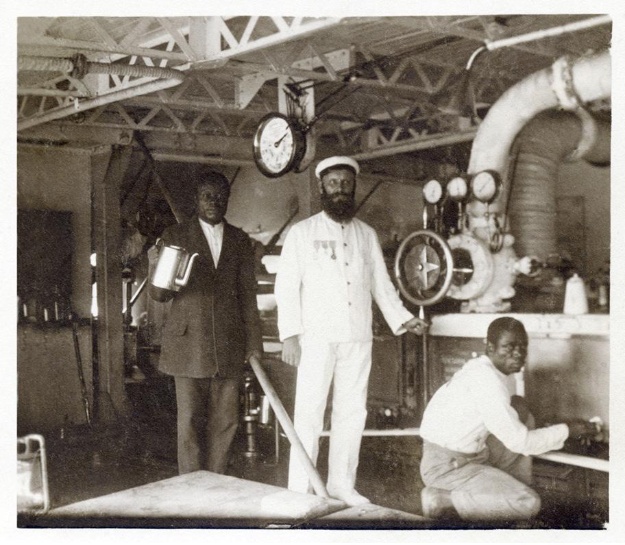 Ruskeansävyinen valokuva laivan konehuoneesta. Kahden tummaihoisen työntekijän keskellä seisoo valkoihoinen mies valkoisessa puvussa ja hatussa.
