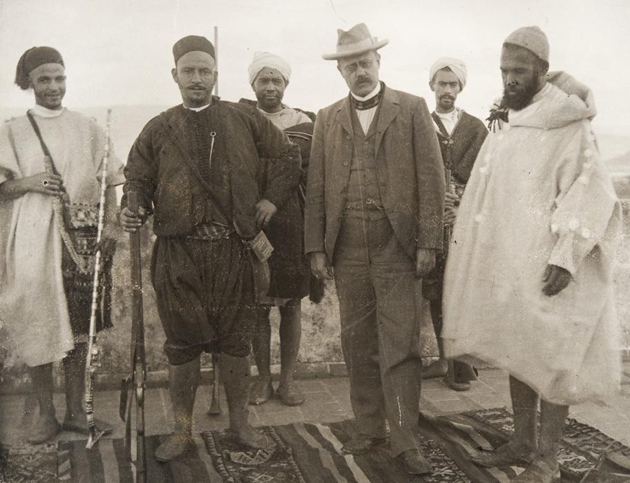 Ruskeansävyinen valokuva ulkona seisovasta kuuden miehen ryhmästä. Muut miehet ovat pukeutuneet erilaisiin paikallisiin asuihin, Edward Westermarck länsimaiseen miesten pukuun, liiviin ja hattuun.