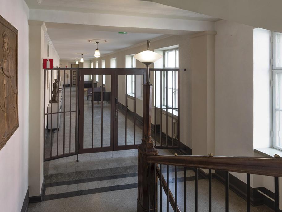 Näkymä portaikosta käytävään. Portaikon ja käytävän välissä on ruskea teräs- ja puurakenteinen portti. Kuvan lähde: Arno de la Chapelle.