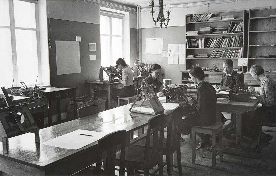 Mustavalkoinen valokuva huoneesta, jossa istuu neljä naista ja yksi mies kirjotuskoneiden ja muiden kojeiden ääressä työskentelemässä.