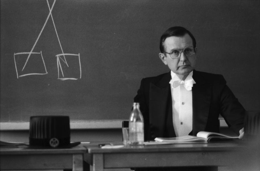 Mustavalkoinen kuva, jossa Matti Klinge istuu hienossa puvussa liitutaulun edessä.