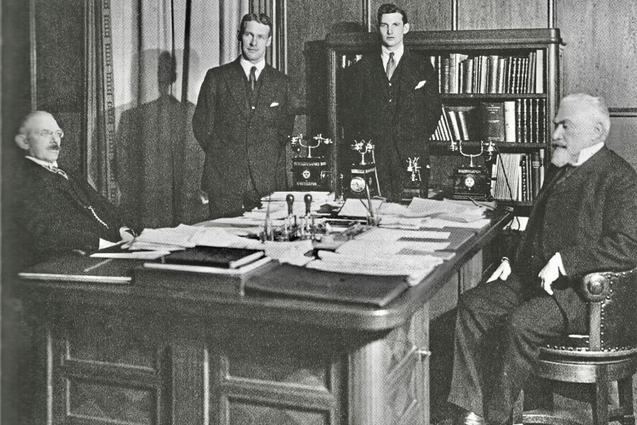 Mustavalkoinen valokuva, jossa neljä tummiin pukuihin pukeutunutta miestä on asettuneena suuren puisen pöydän ääreen. Kaksi vanhempaa miestä istuu, keskellä olevat kaksi nuorempaa miestä seisovat. Pöydällä on paljon papereita ja useampi puhelin.
