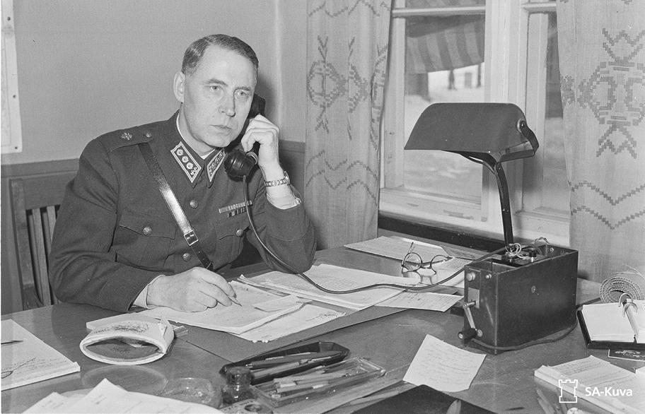 Mustavalkoinen valokuva, jossa sotilaspukuinen mies istuu työpöydän ääressä ja phuu puhelimeen.