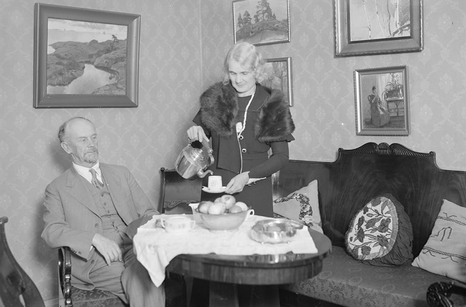 Mustavalkoinen valokuva iäkkäästä pariskunnasta tauluin koristellussa huoneessa. Nainen kaataa kahvia, mies istuu kahvipöydän ääressä.