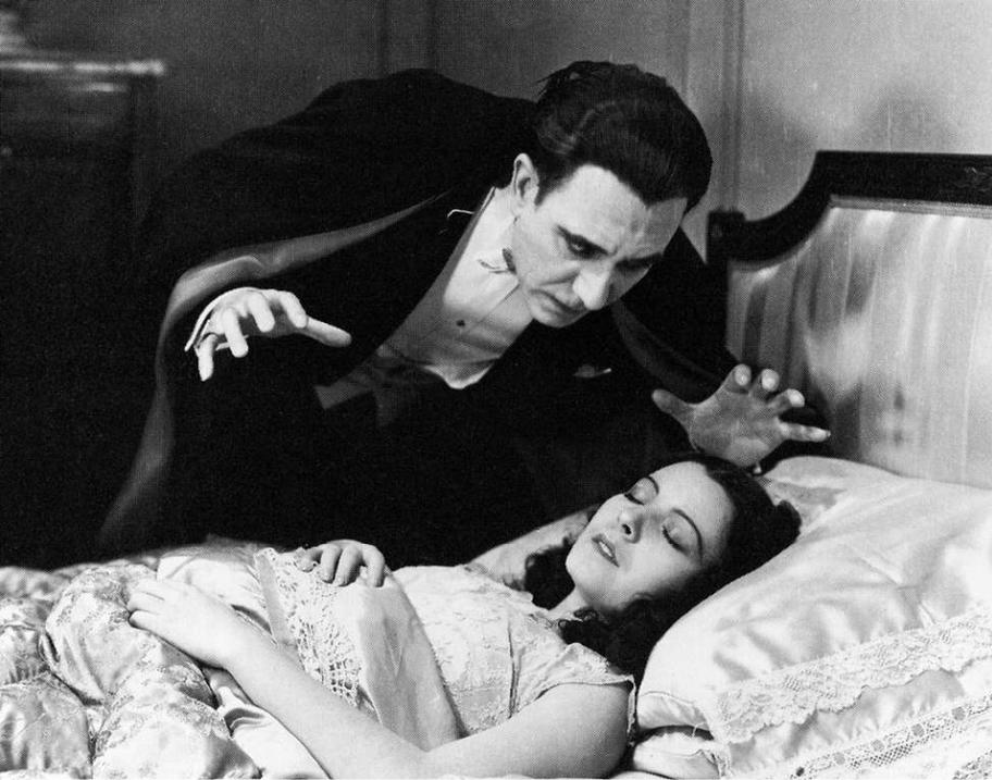 Mustavalkoinen valokuva, jossa pukuun ja mustaan viittaan pukeutunut mies on kumartuneena nukkuvan naisen ylle.