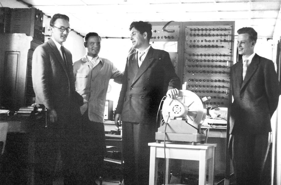 Mustavalkoinen valokuva, jossa neljä miestä puvuissa seisoo erilaisten laitteiden äärellä.