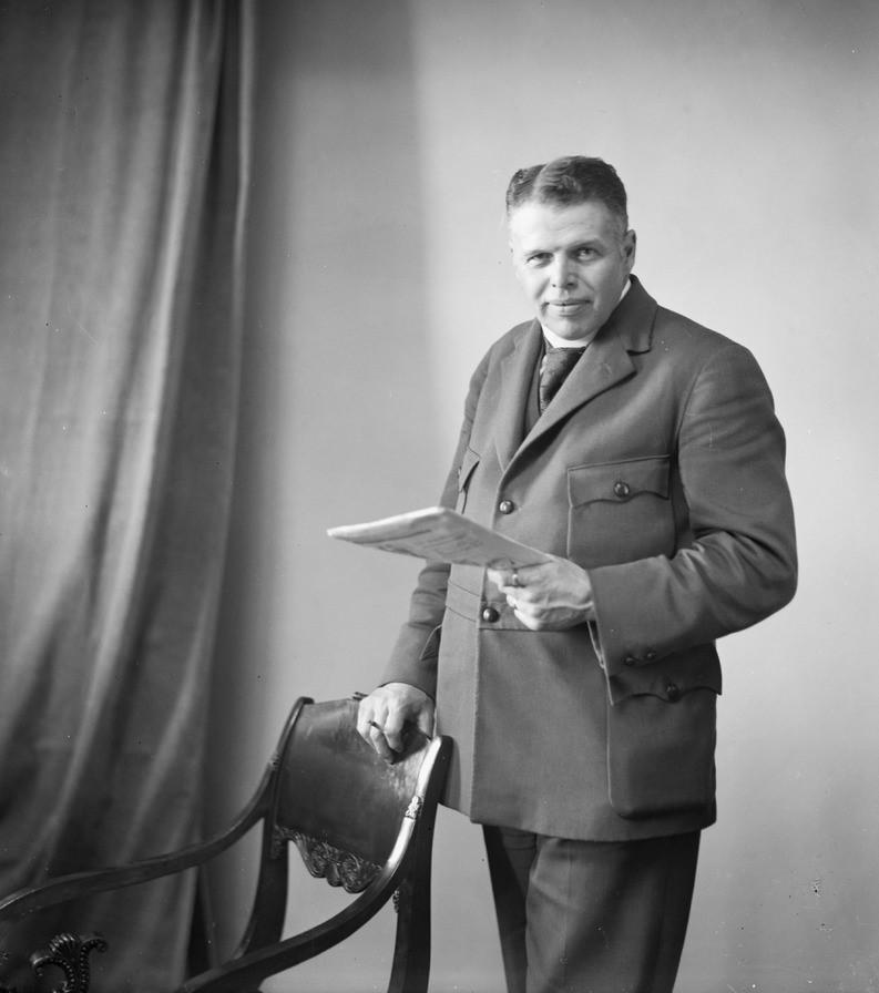 Mustavalkoinen kuva Carl-Gustaf Herlitzistä, joka seisoo pidellen toista kättään tuolin selkänojalla.