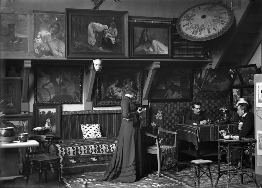 Mustavalkoinen kuva korkeasta huoneesta, jonka seinillä on paljon tauluja. Nainen kävelee huoneen keskellä. Huoneen reunassa on kaksi miestä, jotka istuvat pöydän ääressä.