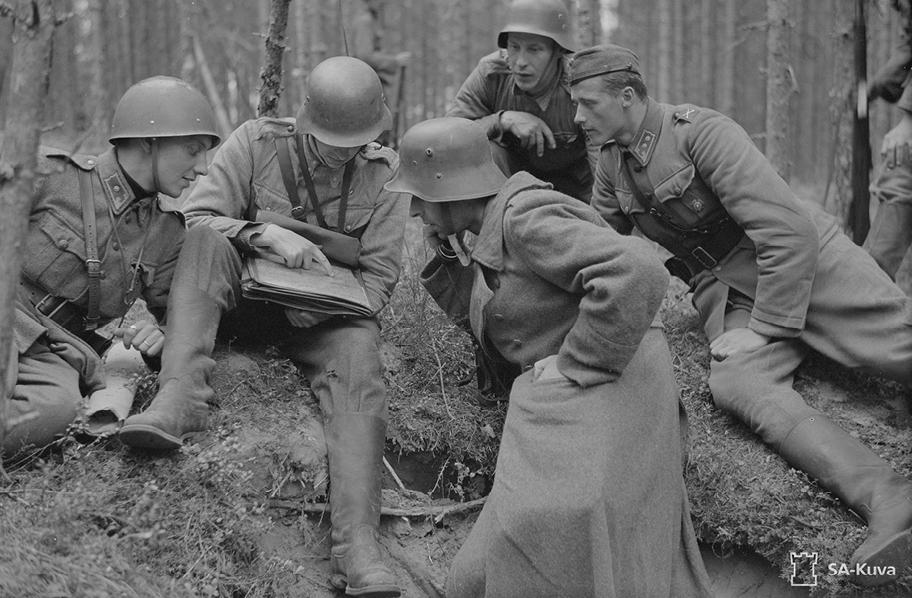 Mustavalkoinen kuva, jossa viisi sotilasta on kumartunut metsässä tutkimaan dokumenttia..
