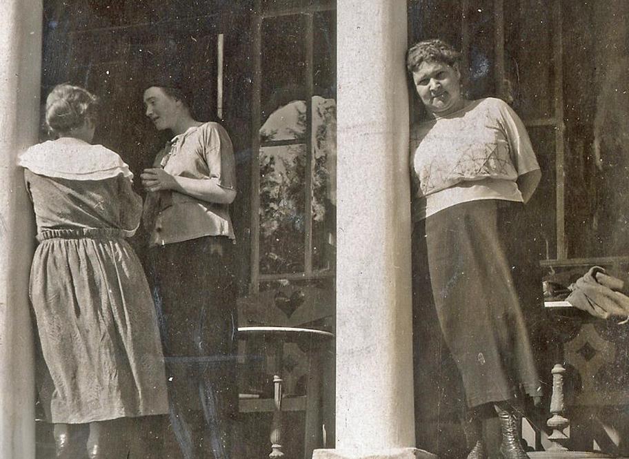 Mustavalkoinen kuva, jossa kolme naista seisoo terassilla. Kaksi heistä on kääntynyt keskustelemaan keskenään. Jenny Markelin nojaa pylvääseen ja katsoo kameraan.
