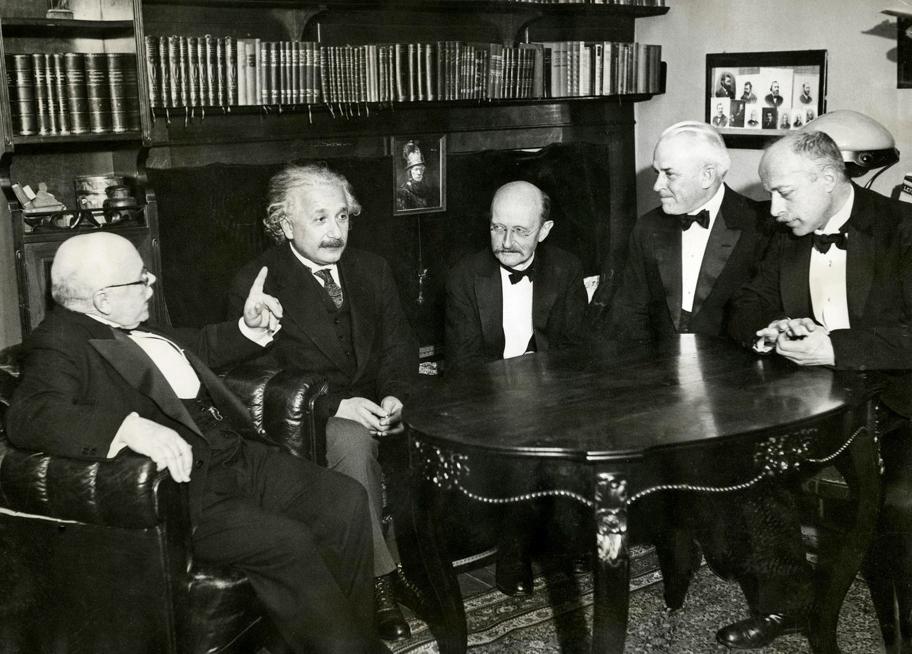 Mustavalkoinen valokuva viidestä miehestä koristeellisen pöydän ääressä. Miehet ovat pukeutuneet tummiin pukuihin ja solmioihin. Taustalla on kirjahylly.