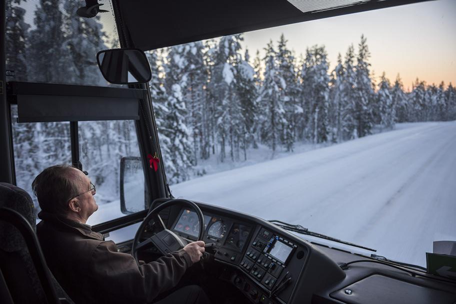 Näkymä linja-auton etuosasta. Kuljettaja ohjaa linja-autoa lumisessa, hämärtyvässä maisemassa.