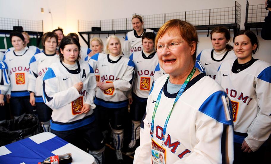 Punatukkainen nainen seisoo pelaajajoukon keskellä iloisen näköisenä. Kaikilla on päällään sinivalkoiset peliasut.