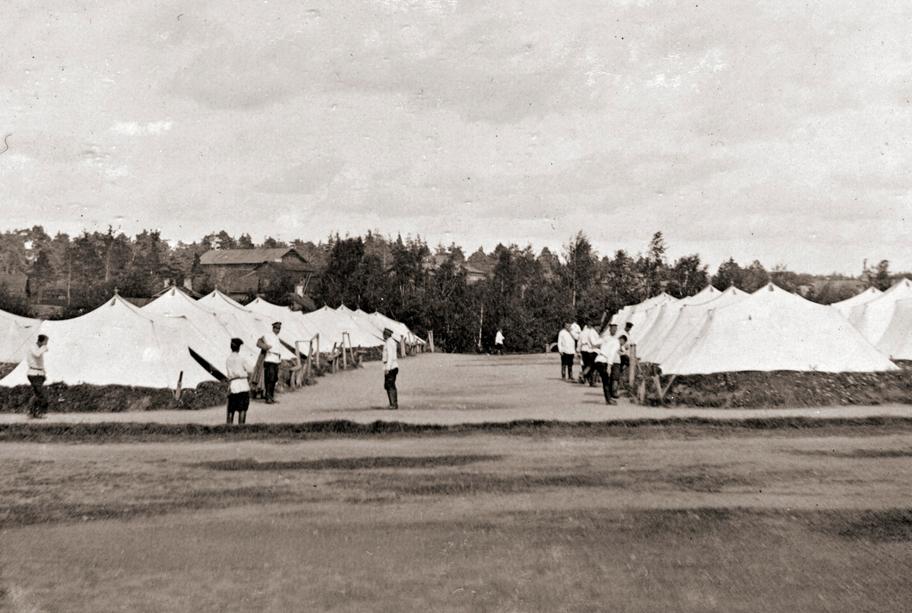 Mustavalkoinen valokuva avaralta kentältä, jolla sijaitsee lukuisia suuria telttoja. Telttojen luona kulkee vajaa kymmenen samanlaisiin asuihin pukeutunutta miestä.