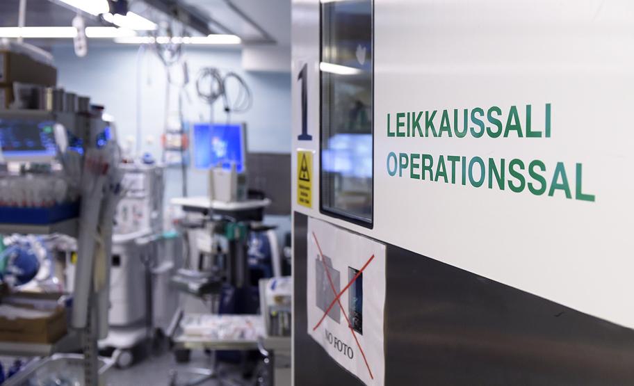 Ovi auki huoneeseen, jossa näkyy runsaasti erilaista tekniikkaa ja välineistöä. Ovessa lukee Leikkaussali −Operationssal.