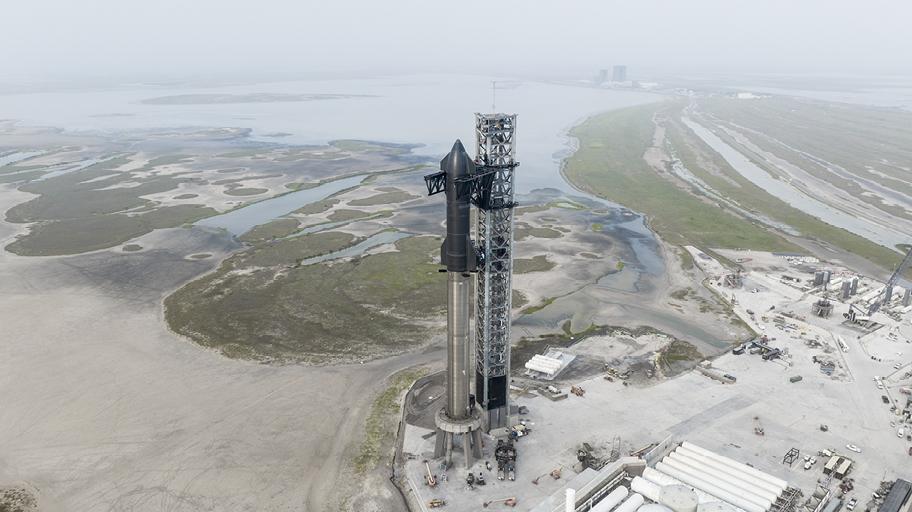 Suuri teline ja siinä kiinni oleva raketti sekä laajempi erilaista tekniikkaa sisältävä alue tasaisessa maisemassa.