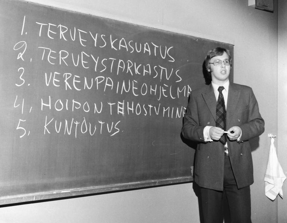Mustavalkoinen valokuva, jossa Kai Koskela seisoo liitutaulun edessä puhumassa. Hänellä on päällään puku ja kädessään liitu. Taululla lukee "1. terveyskasvatus 2. terveystarkastus 3. verenpaineohjelma 4. hoidon tehostaminen 5. kuntoutus".