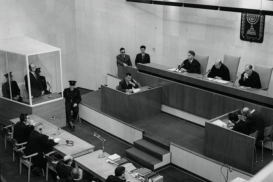 Mustavalkoinen kuva oikeussalista, jossa syytetty Adolf Eichmann seisoo vasemmalla lasitetulla paikalla.