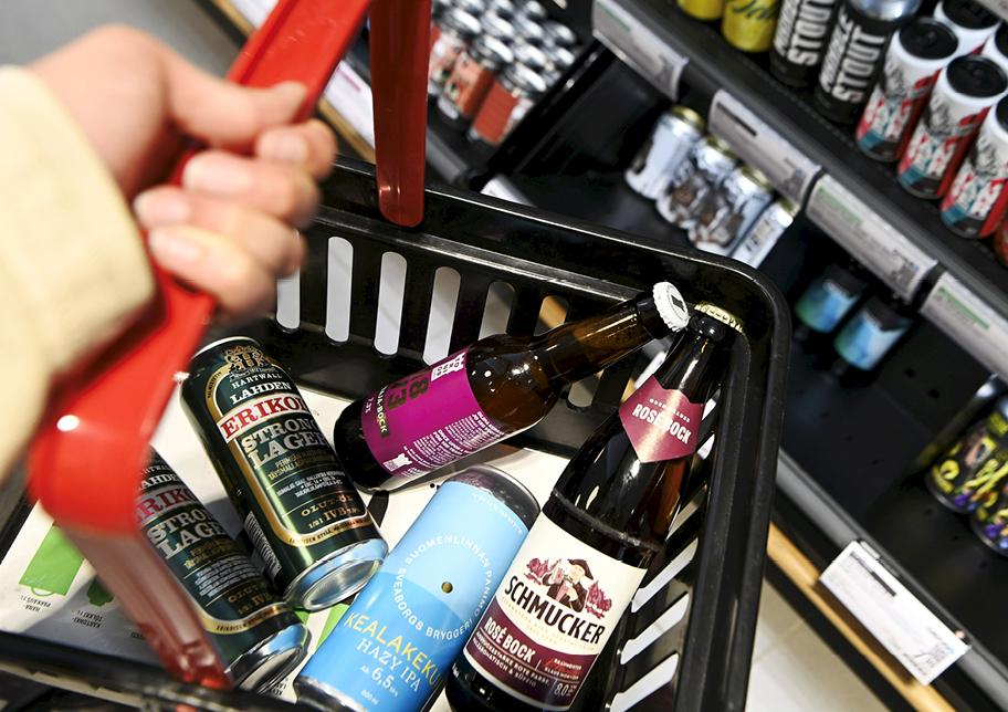Ostoskori, jossa on kolme tölkkiä ja kaksi pulloa erimerkkisiä olluita. Taustalla näkyy hylly, jossa on runsaasti lisää alkoholijuomia.