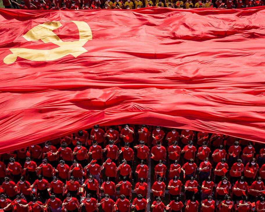 Punaisiin pukeutuneita nuoria seisoo katsomossa. Heidän päälleen on avautumassa suuri kankainen Kiinan lippu.