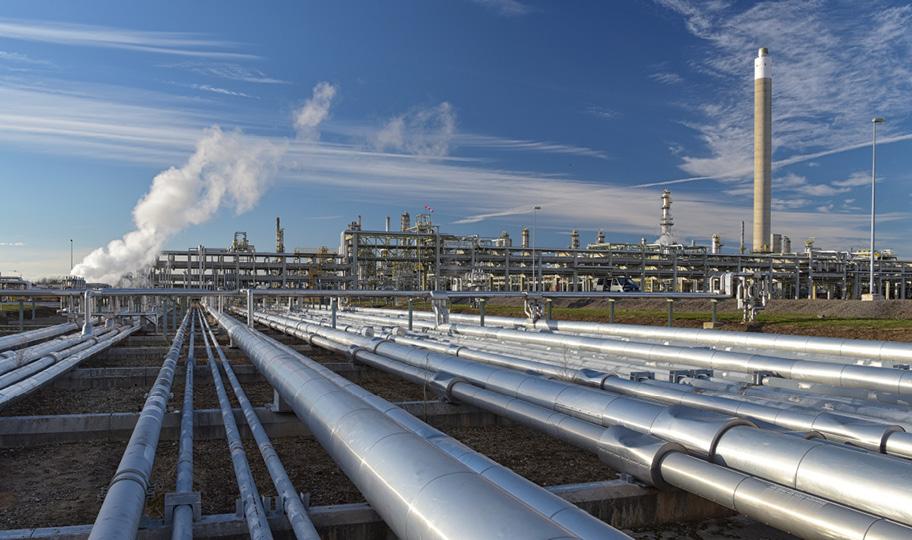 Maisemakuva, jossa näkyy suuri teollisuuslaitos. Etualalla kulkee paljon metallisia putkia, taustalla näkyy hieman savua tai vesihöyryä ja korkea piippu.