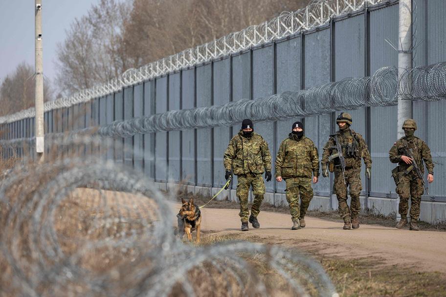 Neljä sotilasasuihin pukeutunutta henkilöä ja saksanpaimenkoira kävelee korkean harmaan aidan vieressä kulkevalla hiekkatiellä. Kahdella henkilöllä on aseet näkyvissä. Aidan päällä ja sivuissa on piikkilankaa, jota näkyy myös kuvan etualalla.