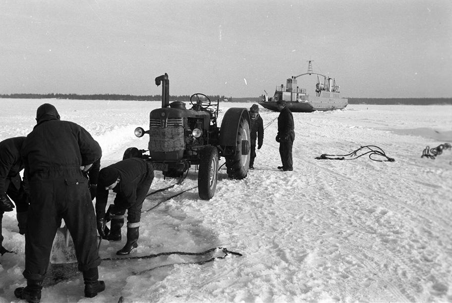 Mustavalkoinen valokuva, jossa on viisi miestä, traktori ja laiva jäätyneellä merellä.