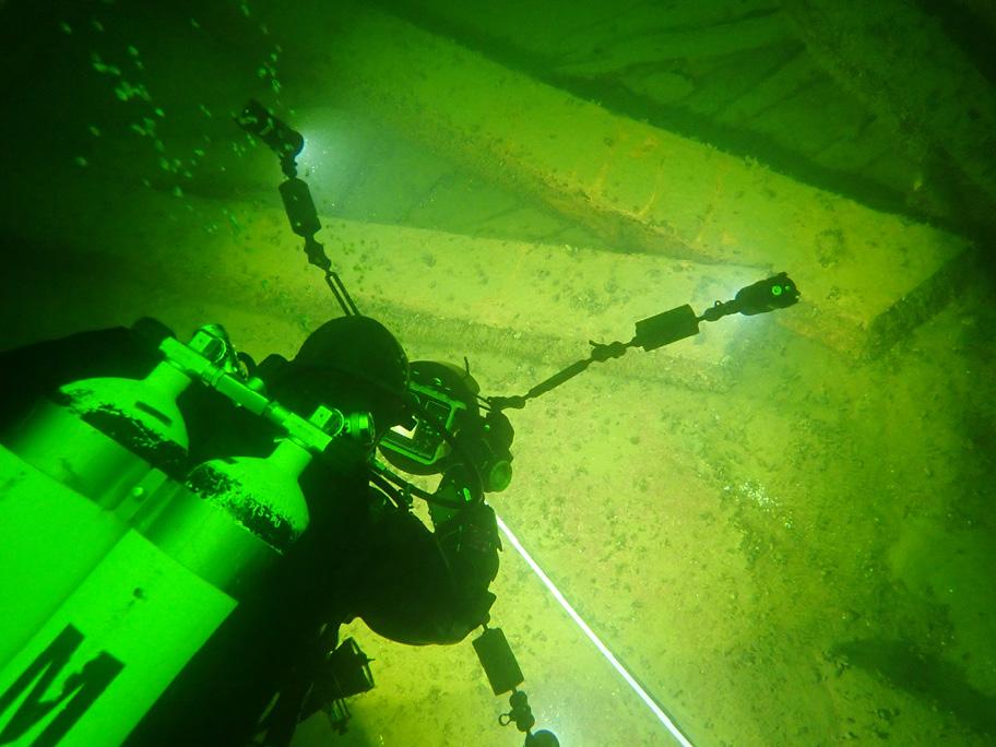 Sukeltaja happipulloineen kuvaa veden alla näkyviä suuria palkin näköisiä elementtejä. Maisema on vihertävä ja kamera valaisee kuvattavaa kohdetta.