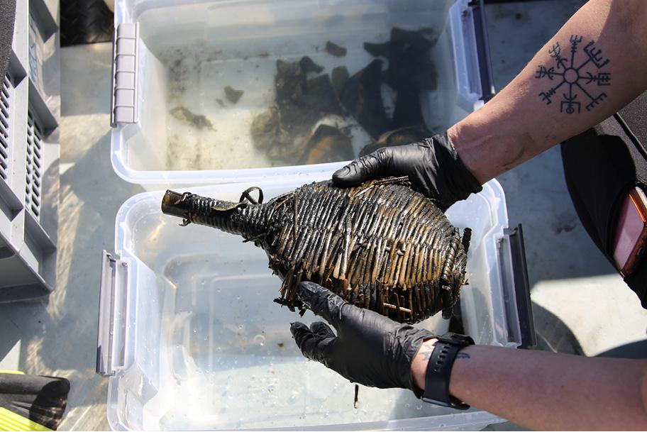 Henkilö pitää käsissään leveämahaista pulloa, jonka päällä on kostean näköinen ja hieman hajonnut puinen punos. Henkilöllä on käsissään kumihanskat ja alla näkyy muovisia astioita, joissa on vettä.