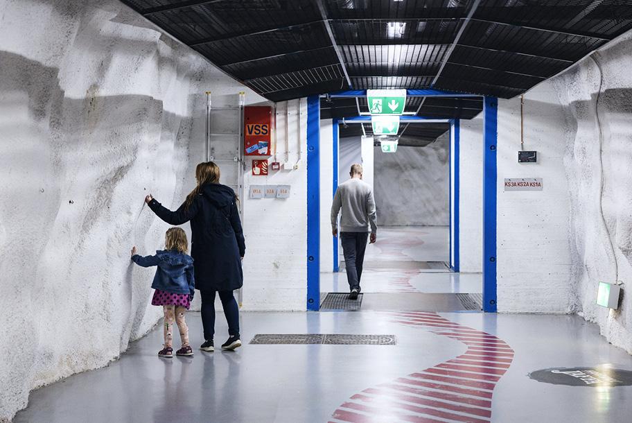 Ikkunaton vaaleaseinäinen leveä käytävä, jossa kävelee muutamia ihmisiä. Seinillä on joitakin laitteita johtoineen.