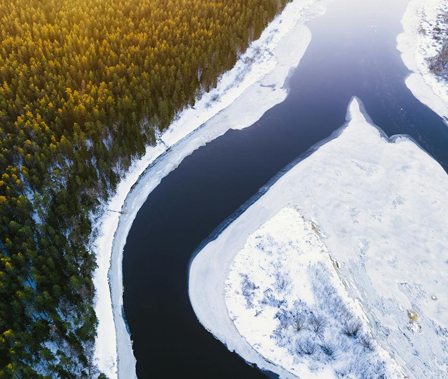 Avara maisema ylhäältä päin kuvattuna. Kahteen haaraan jakautuva joki halkoo tiheää metsää. Joen keskellä on saari. Joen rannat ja saari ovat jään ja lumen peitossa.