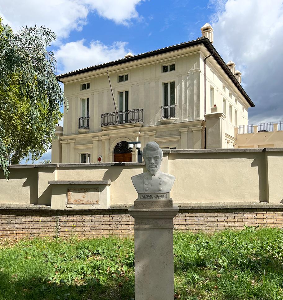 Vaalea kivitalo. Talon edessä on aita ja aidan edessä vaaleasta kivestä tehty miehen rintakuva. Talon vieressä on tuuhea lehtipuu.