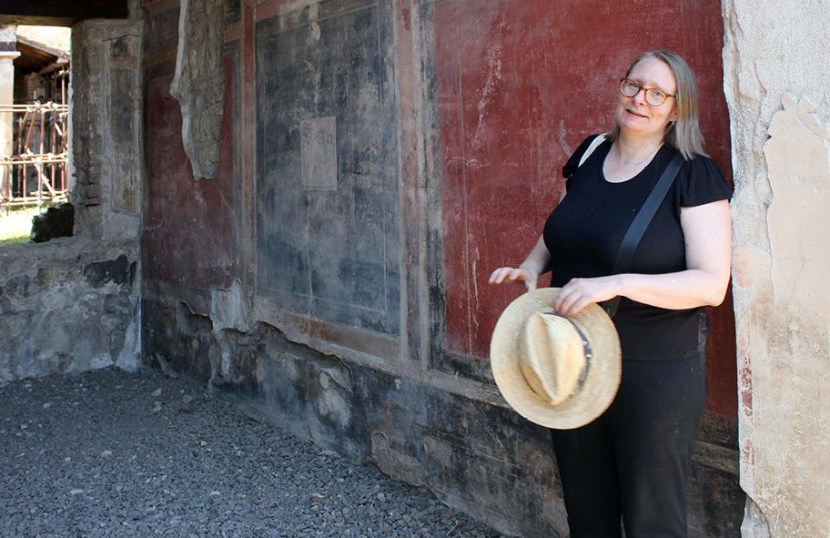 Naisoletettu seisoo kivitalon seinän edessä, kädessään olkihattu. Seinä ja sen punainen maali ovat kuluneita.