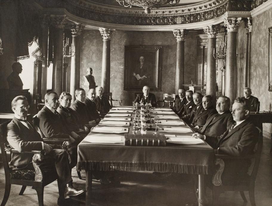 Mustavalkoinen valokuva koristeellisesta salista, jossa istuu neljätoista henkilöä pitkän pöydän ääressä.