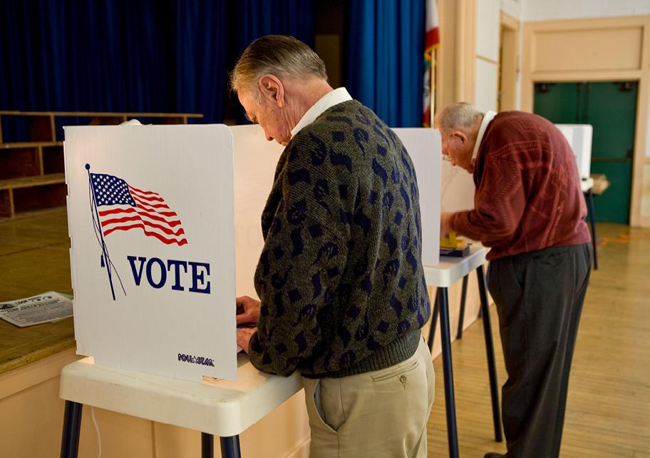 Muutamia äänestyskoppeja koulun liikuntasalin näköisessä tilassa. Lähimmän äänestyskopin valkoisessa seinäkkeessä on Yhdysvaltain lipun kuva ja teksti "Vote". Kaksi iäkkäämpää miesoletettua on juuri äänestämässä.