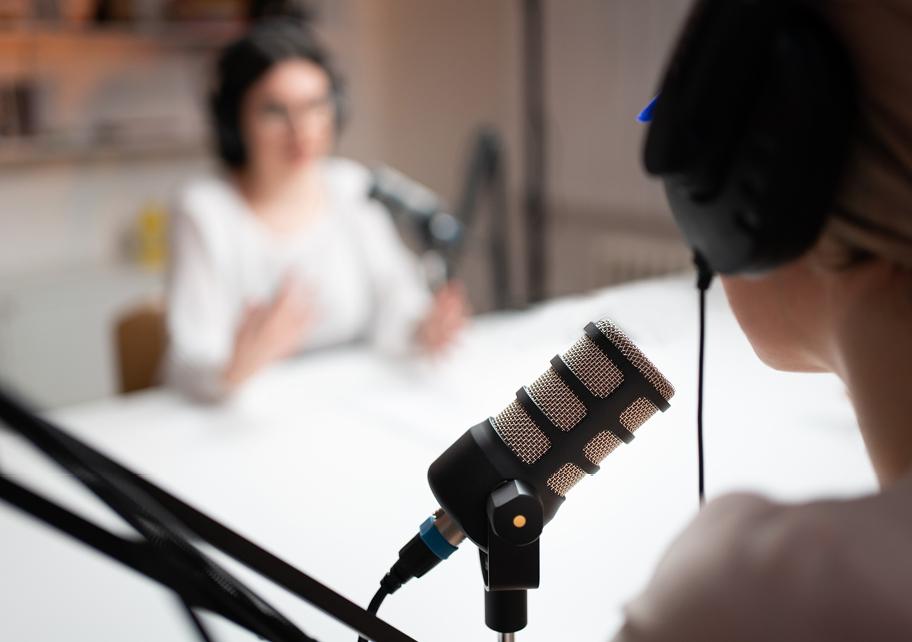 Kaksi henkilöä tekemässä puheohjelmaa. He istuvat pöydän ääressä vastapäätä toisiaan, molemmilla on korvillaan kuulokkeet ja edessään mikrofoni.
