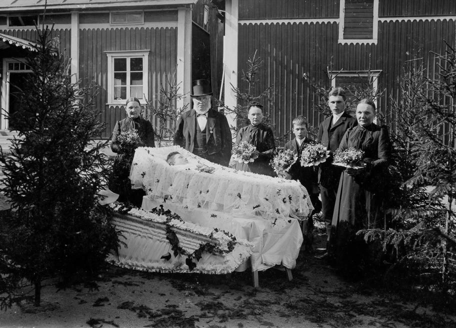 Mustavalkoinen valokuva, jossa kuusi ihmistä seisoo mustissa vaatteissa talon pihamaalla avoimen arkun edessä. Monilla heistä on kukkakimput käsissään. Arkku on vuorattu ja päällystetty valkoisella kankaalla ja arkussa makaa nuori ihminen.