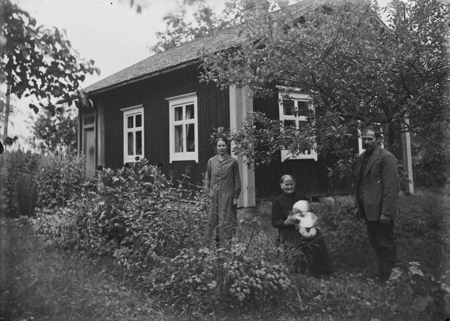 Mustavalkoinen valokuva, jossa on kolme henkilöä puutarhassa puutalon edessä. Nainen ja mies seisovat ja heidän välissään istuu vanhempi nainen vauva sylissään.