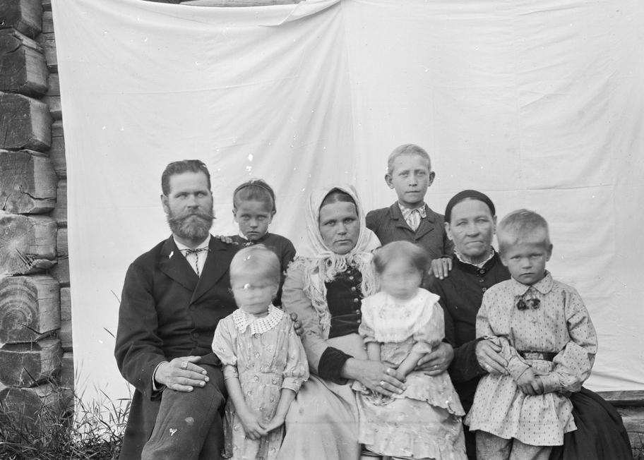 Mustavalkoinen valokuva, jossa perhe poseeraa hirsitalon seinään ripustetun valkoisen kankaan edessä. Ryhmässä on mies ja nainen sekä vanhempi nainen ja viisi eri-ikäistä, melko nuorta lasta.