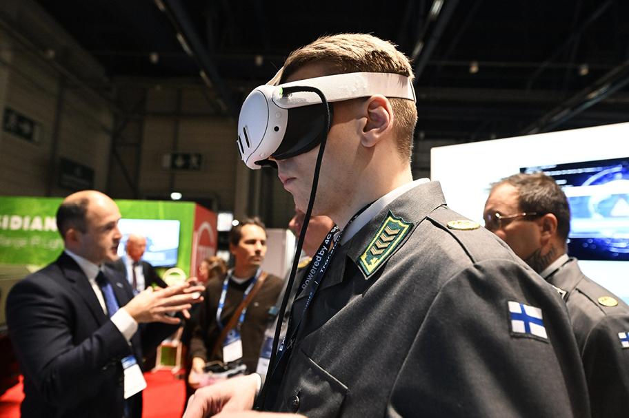 Sotilasasuun pukeutunut henkilö jonkinlaiset virtuaalitodellisuuteen liittyvät lasit päässään. Taustalla näkyy messuvieraita.